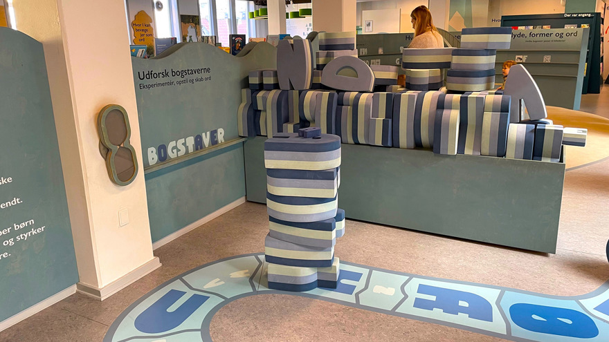 Ett biblioteksrum som går i färgerna grått, blått och beige. På golvet finns en målad gångväg med bokstäver, stora bokstäver i ett lätt material ligger i högar och staplar för att kunna leka med. På en vägg står det Utforska bokstäverna på danska.