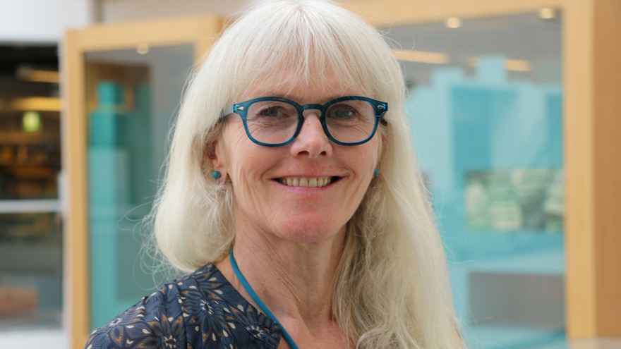 Jeanette Pejtersen är i övre medelåldern. Hon har långt vitt hår, bär ett par glasögon med blå bågar och en mönstrad blus. Hon ler mot fotografen, hon står i en inomhusmiljö.