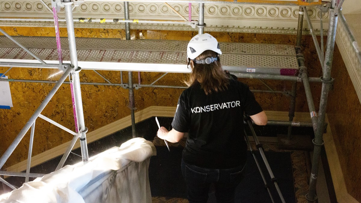 I ett dekorerat trapphus med byggnadsställningar står en person med vit bygghjälm och en t-shirt med texten Konservator på ryggen.