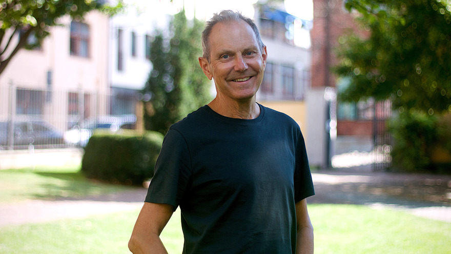 Tomas Tengby har grått kortklippt hår som är kammat bakåt. Han är klädd i en svart t-shirt och står utomhus. Han står i stadsmiljö, bakom honom finns en gata med hus, stora träd och en grind in till en asfalterad gård. Han ler på bilden och tittar rakt mot betraktaren.