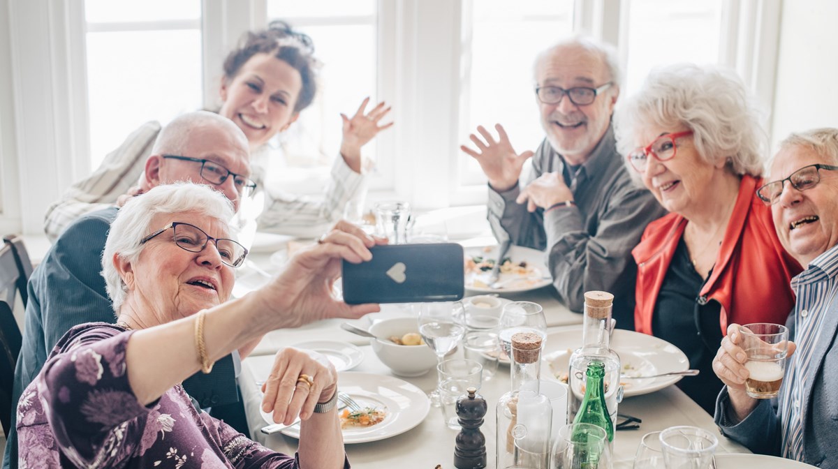 Glada seniorer vid ett matbord. De vinkar och en person tar ett foto av dem alla med sin mobil.