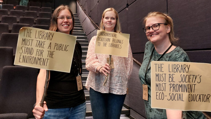 Johanna Juvonen, Tuula Rönkä,och Liisa Uimonen står med varsitt plakat. samtliga plakat har budskap på engelska som handlar om att biblioteket måste agera för klimatet. De tre kvinnorna är alla i 30-35-årsåldern och de står i en trappa i en lokal med grå väggar och grå rader med sittplatser.