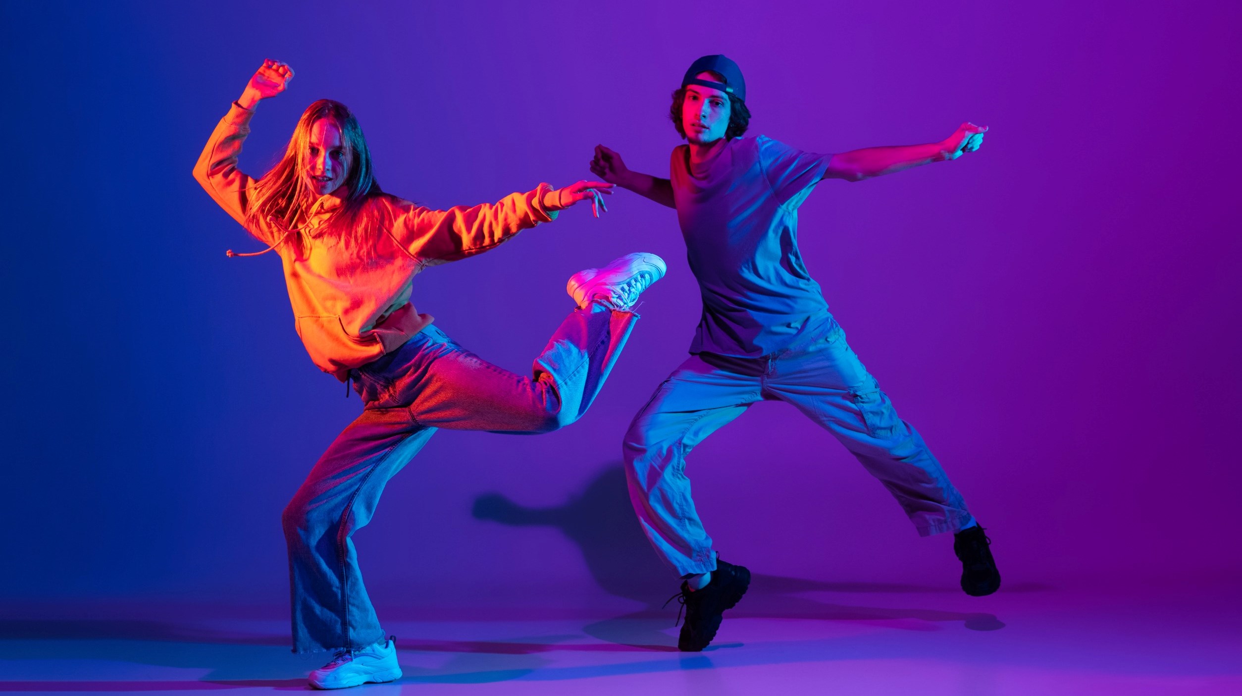 Young man and woman dancing hip-hop in casual sports youth clothes on gradient purple pink background at dance hall in neon light.