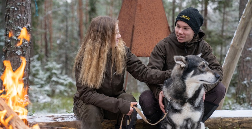 Två elever sitter tillsammans med en hund framför en brasa i skogen