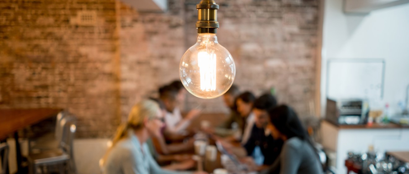 Bild med en lysande glödlampa i förgrunden. Bakgrunden är suddig och visar en grupp människor som sitter och arbetar tillsammans runt ett bord.