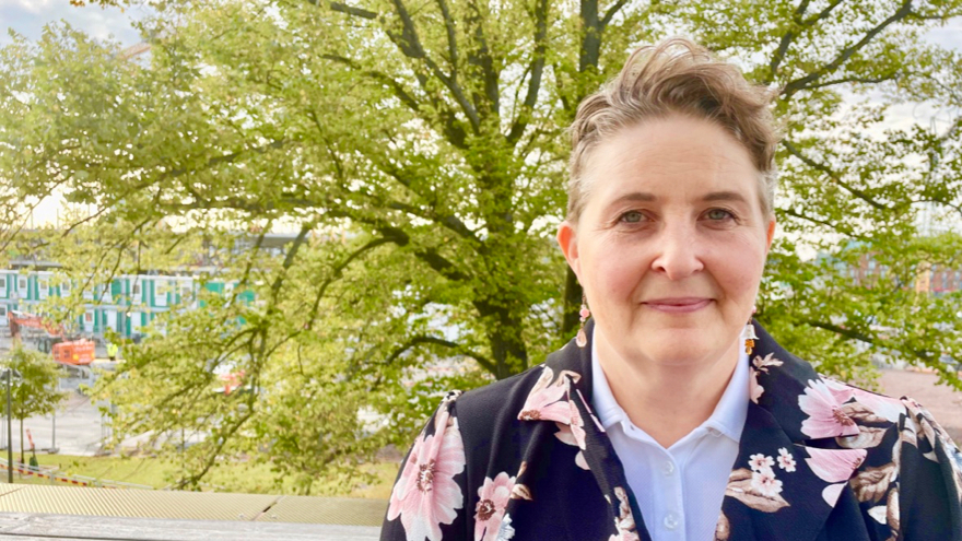 Åsa-Maria Levinsson Berg har kortklippt ljusbrunt hår. Hon är klädd i en vit blus och en blommönstrad kavaj. I bakgrunden finns ett grönskande träd.