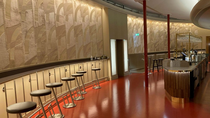 Den nyrenoverade foajébaren på Stadsteatern i Göteborg. en böjd fondvägg klädd med den gyllene tapeten med geometriskt mönster. Utmed väggen löper en väggfast smal borsyta med barstolar placerade med jämna intervall. Till höger i rummet en bardisk klädd i trä. Golvet är rött och högblankt.