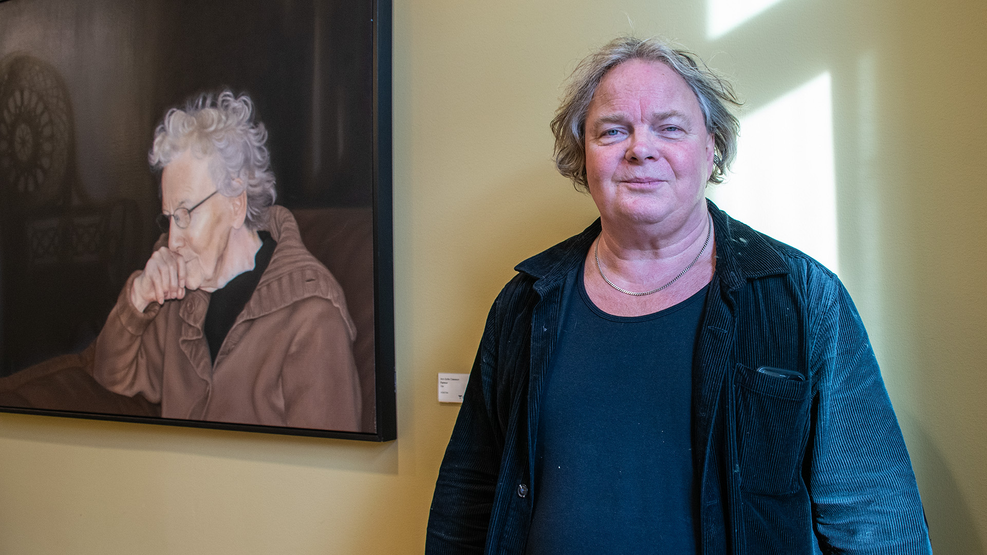 Magnus William-Olsson är 65-årsåldern, han har gråsprängt lite lockigt hår och är klädd i en svart t-shirt och en manchesterjacka. Han står mot en gul vägg, bredvid honom hänger en tavla med en gammal kvinna på. Solen från fönstret gör avspeglingar på väggen bakom honom.