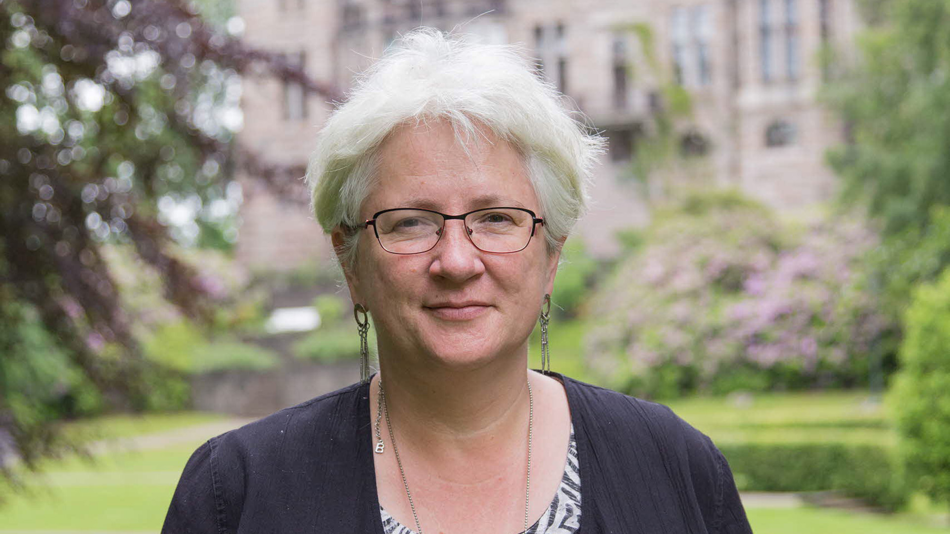 Maria Nilson är i 60-årsåldern. Hon har vitt kortklippt hår, bär glasögon och stora örhängen i silver. Hon står utomhus, i den suddiga bakgrunden finns en byggnad, grönska och blommande buskar. Hon är klädd i en svart blus, tittar rakt in i kameran och ler med stängd mun.