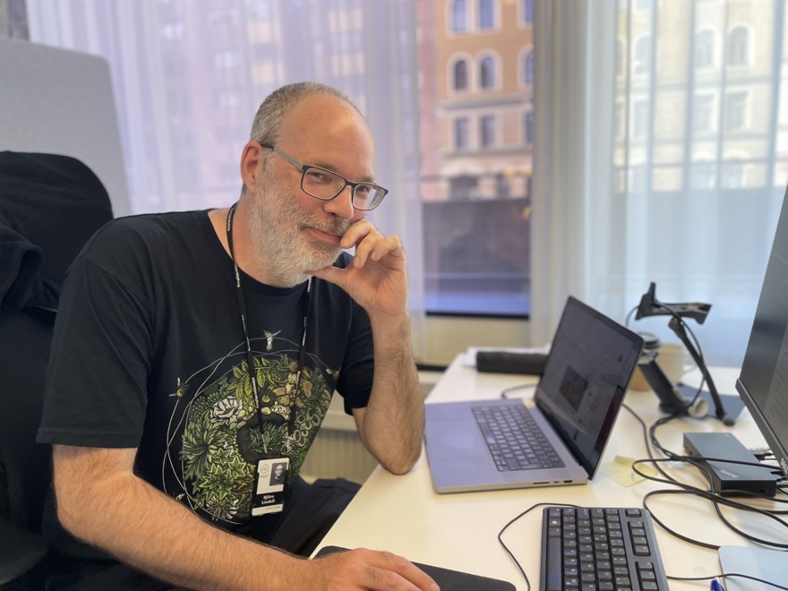 Björn Lindell sitter vid ett skrivbord, framför sig har han en laptop. Han är i 50-årsåldern, har skägg och glasögon och bär en svart t-shirt med tryck.