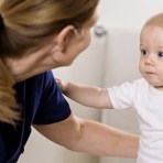 En kvinna i blå läkarrock tittar på ett litet barn som bär en vit body. Vi ser kvinnans rygg och hästsvans och barnet framifrån. Barnet sitter själv men kvinnan stöttar med sin arm bakom barnets rygg.