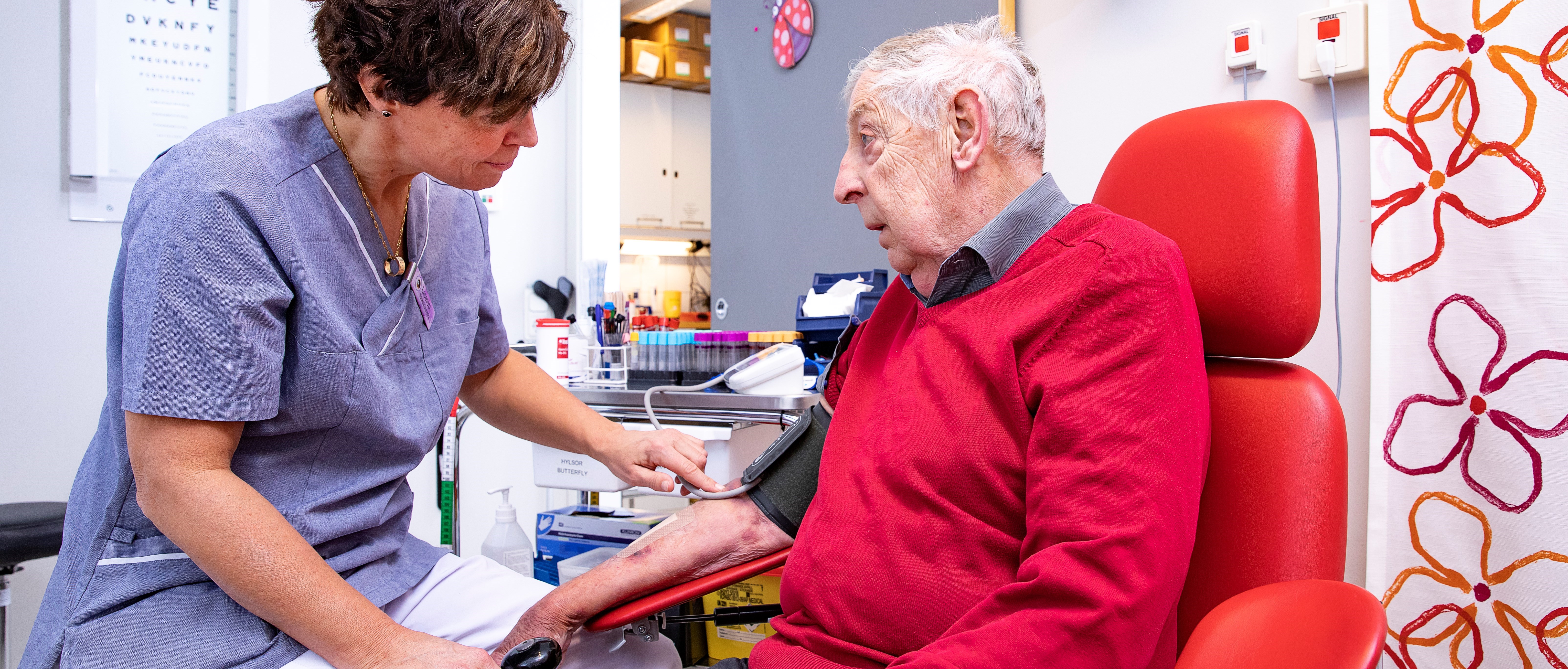 En kvinna i vårdklädsel tar blodtrycket på en äldre man.