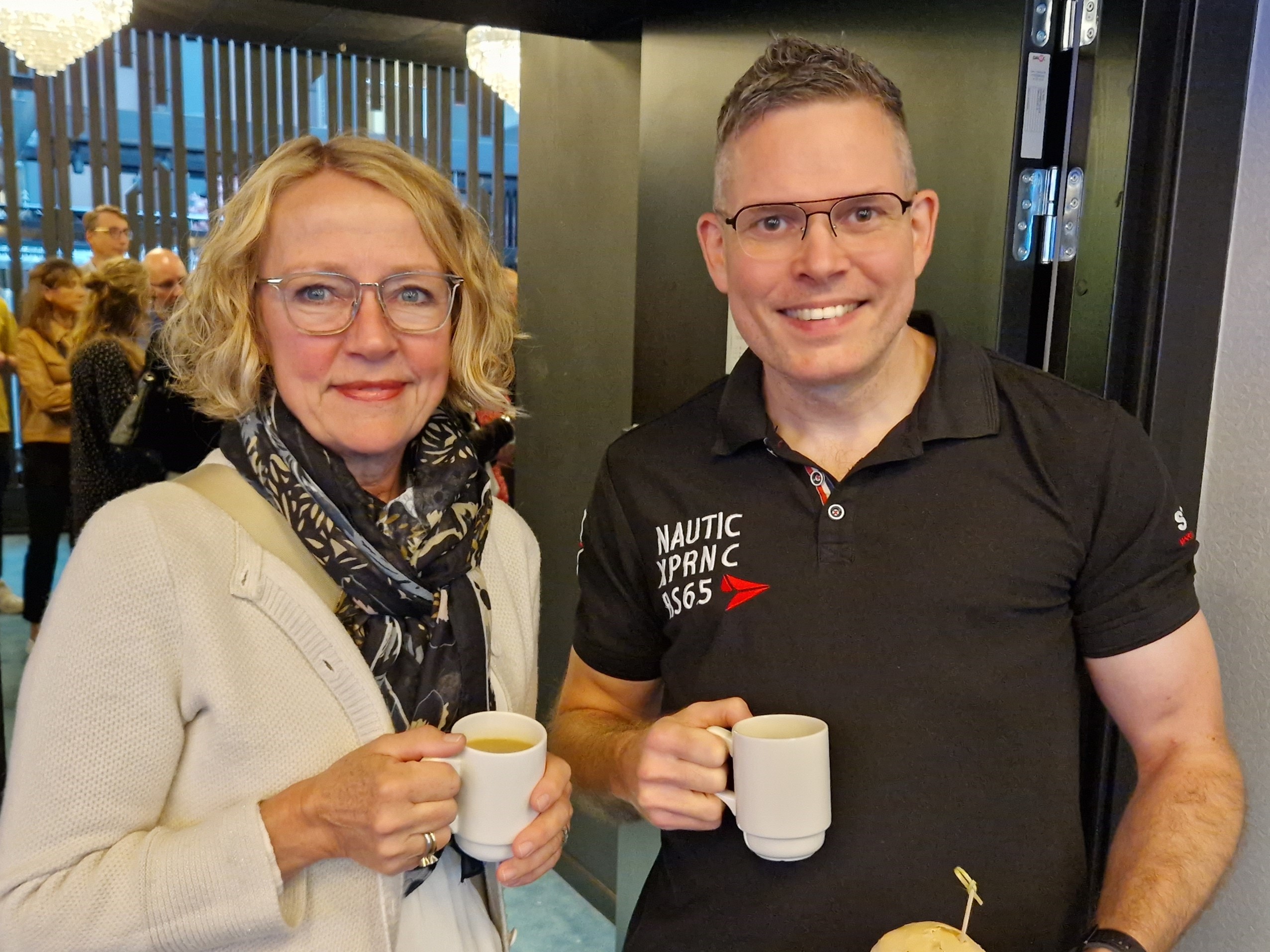 En kvinna och en man står med varsin kopp kaffe i ett större konferensrum.