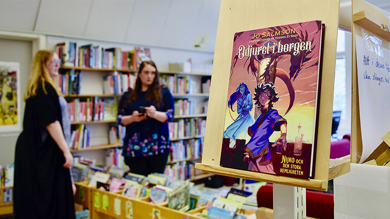 En fantasybok är in-zoomad med kameran. I den lite suddoga bakgrunden står skolbibliotekarerna Elin och Emmy och pratar över en låg bokhylla.