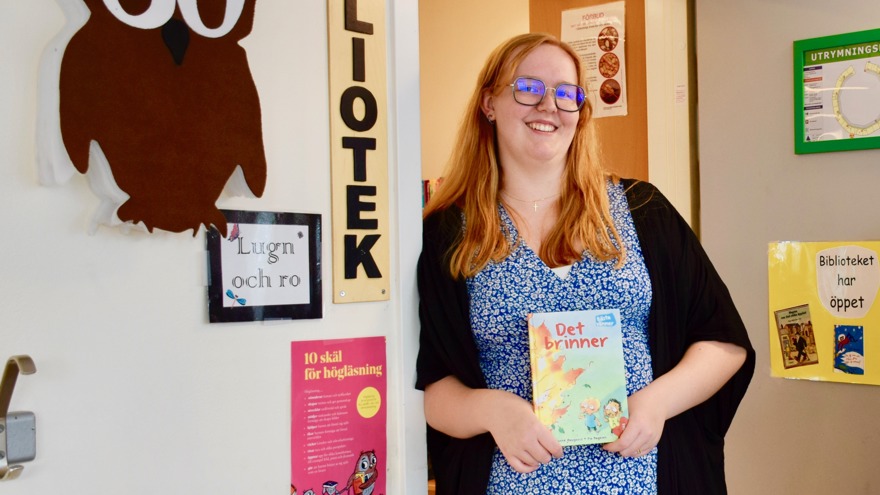 Elin har en blå- och vitmönstrad klänning på sig. Hon har långt blont hår och bär glasögon. Hon står lutad mot en dörrpost, på dörren står ett anslag med meningen "Biblioteket har öppet".