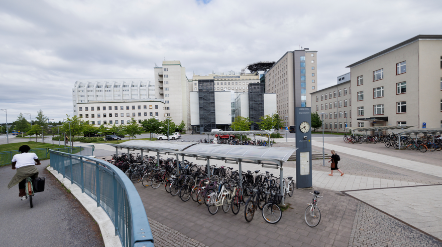 Norrlands universitetssjukhus i Umeå. Stora byggnader i vitt och beige, det är många fönster på husen. Framför dem finns ett cykelställ fyllt med cyklar. Till vänster i bild kommer en cyklist på en cykelbana. Det är molnigt ute och på en hållplatsskylt sitter en klocka som visar tiden.