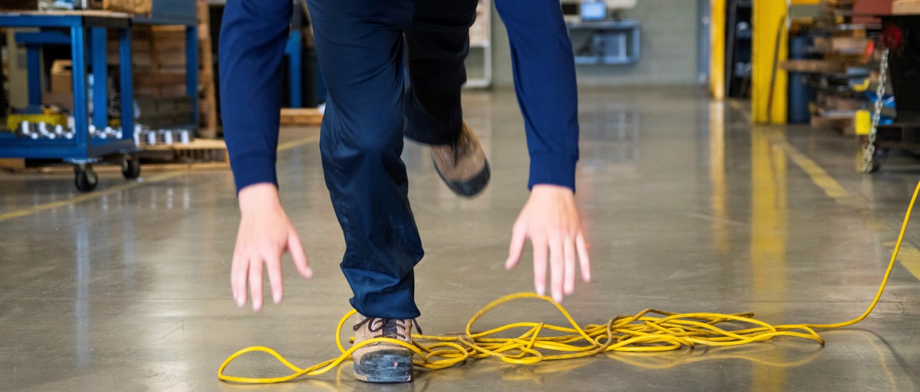 Närbild ur grodperspektiv av en person som snubblar på en sladd på golvet i en verkstad.