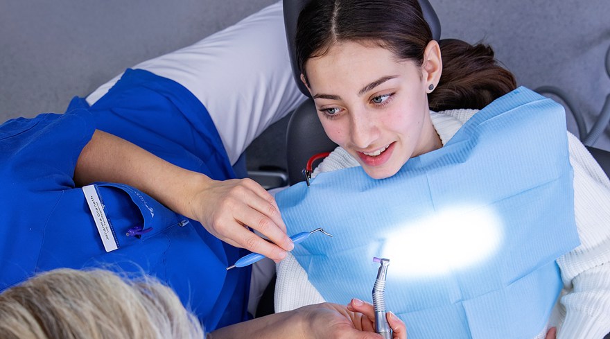 En ung kvinna med mörkt hår sitter i en tandläkarstol bredvid en kortklippt tandhygienist som håller i ett redskap.