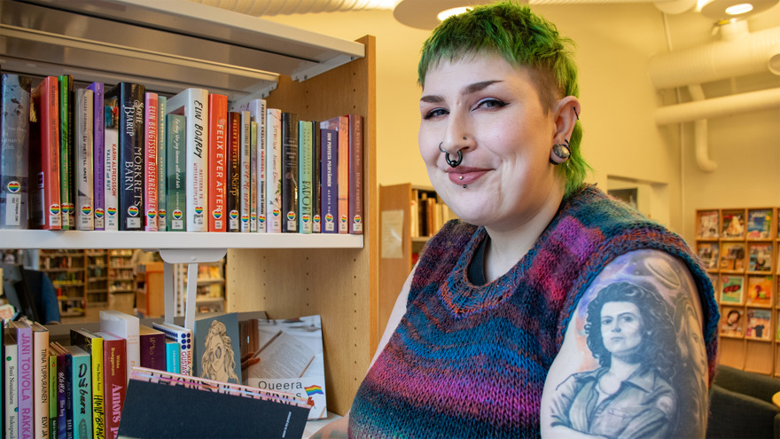 Mikhe står framför en bokhylla i biblioteket. Hen har grönt kortklippt hår och piercingar i bland annat näsa och öron. Hon är klädd i en stickad tröja med korta ärmar, på hennes arm finns en tatuering av skådespelerskan Sigourney Weaver i sin karaktär från filmen Alien. Mikhe tittar in i kameran och ler.