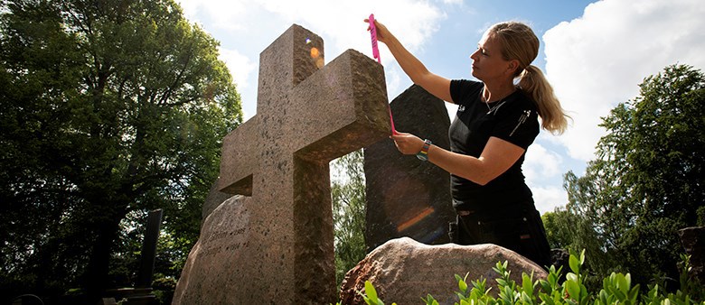 En kvinna står utomhus i strålande sol och mäter upp ett stenkors.Bakom henne skymtar blå himmel och gröna träd.