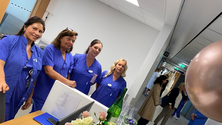Balong och blommor i förgrunden. Fyra blåklädda sjuk- och undersköterskor. Minglande personer i korridor i bakgrunden.
