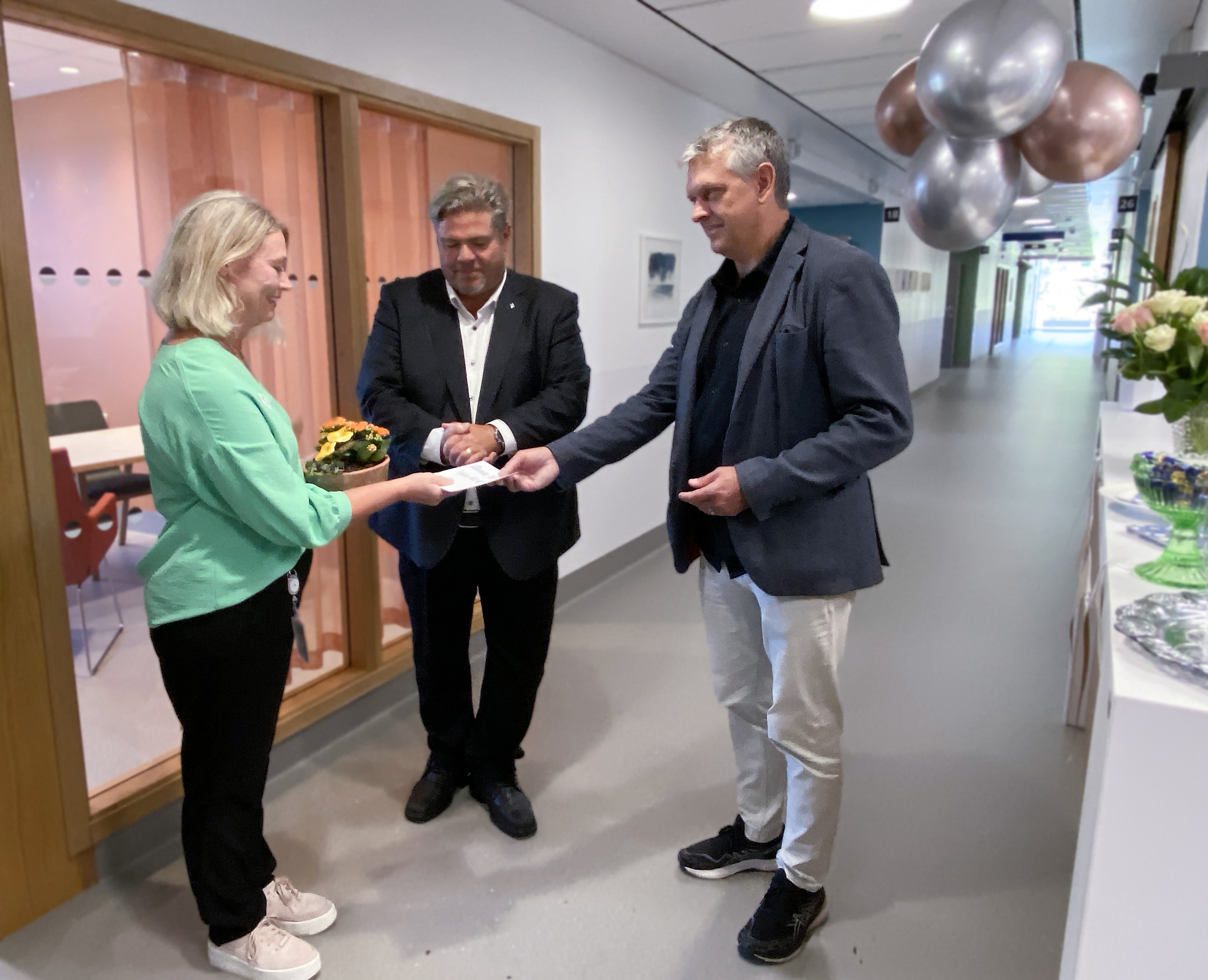 Enhetschef i grön blus får ta emot blomma och kort från Sjukhusen i västers styrelseordförande. Med på bilden också kommunstyrelsens ordförande. Ballonger hänger i taket