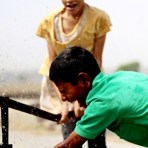 Två barn leker vid en vattenpump, vattnet sprutar och barnen skrattar.