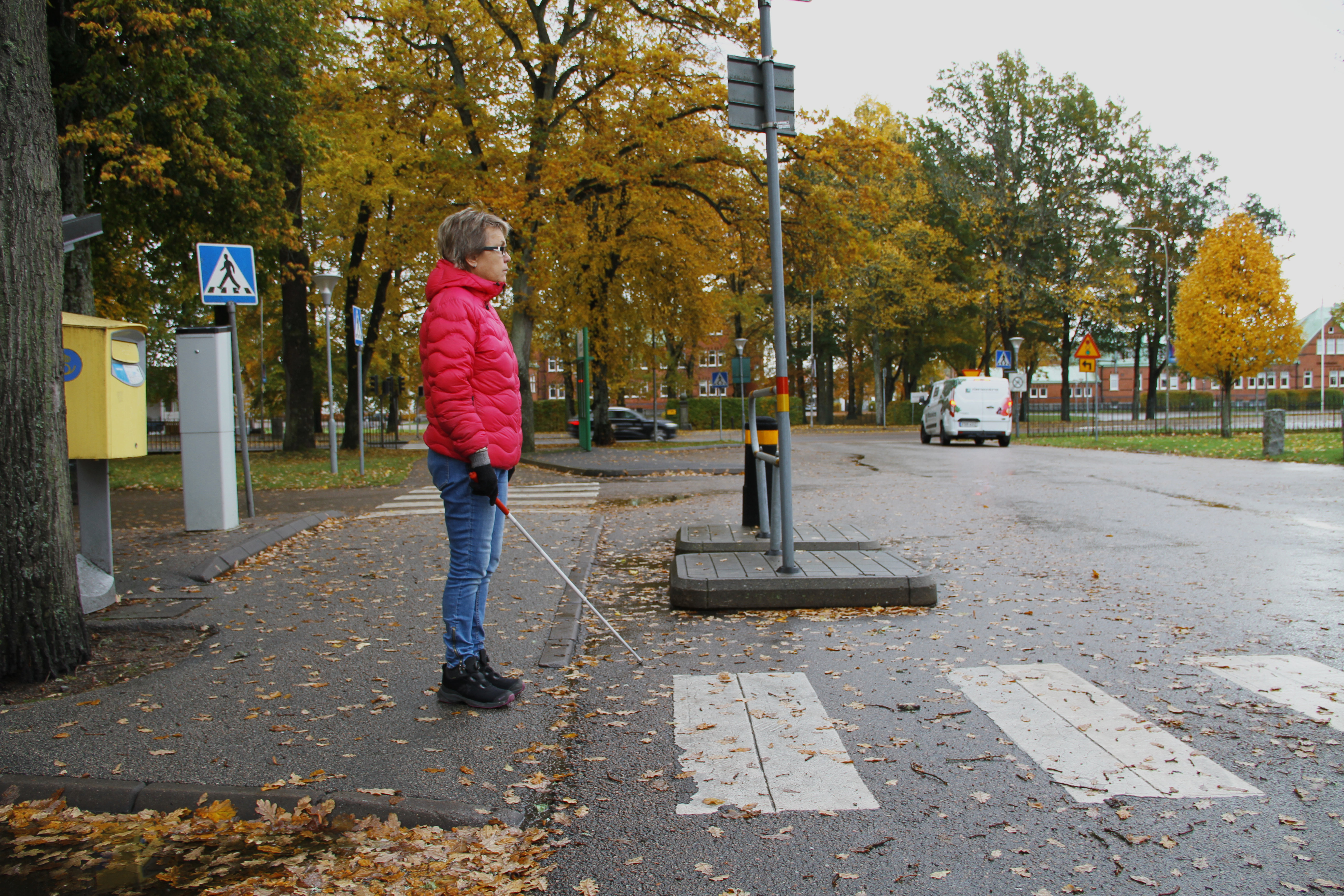 Kvinna med rosa jacka står vid övergångsställe med en käpp framför sig.