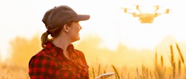 Kvinna med keps och rutig skjorta står i vetefält och tittar upp mot drönare