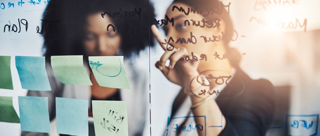 Bild av en glasruta med många anteckningar och lappar i förgrunden, bakom glasrutan står två personer och diskuterar innehållet på glaset.