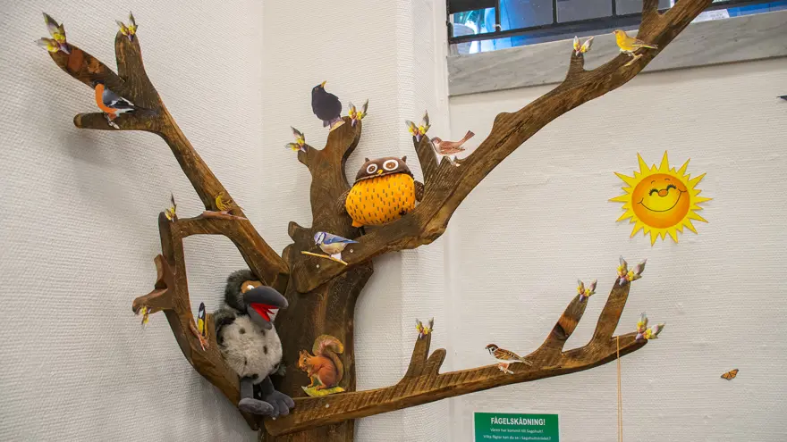 Ett stiliserat träd i hörnet av rummet. I trädet sitter bland annat två gosedjur, en uggla och en kråka. Flera bilder på olika fåglar finns på grenarna.