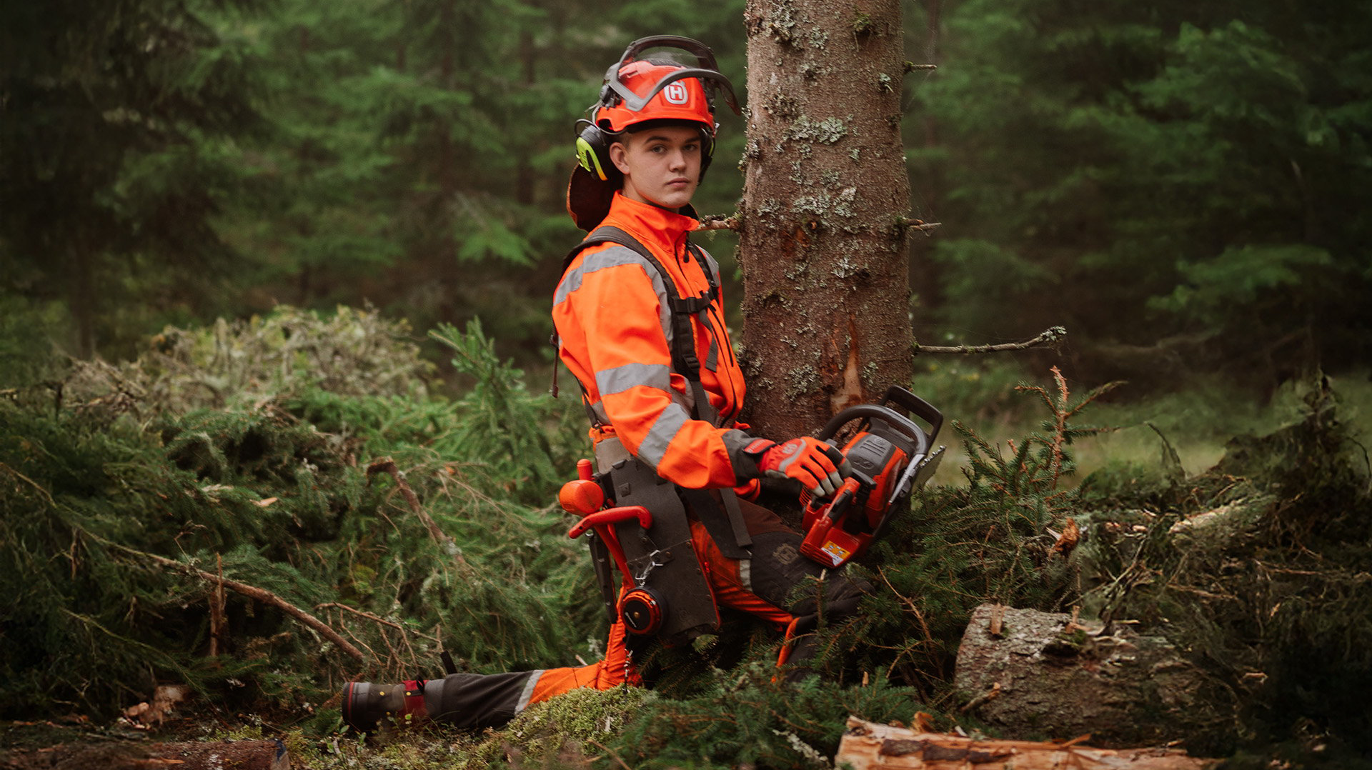 A student wearing protective clothing and a helmet is sitting down, holding a chainsaw
