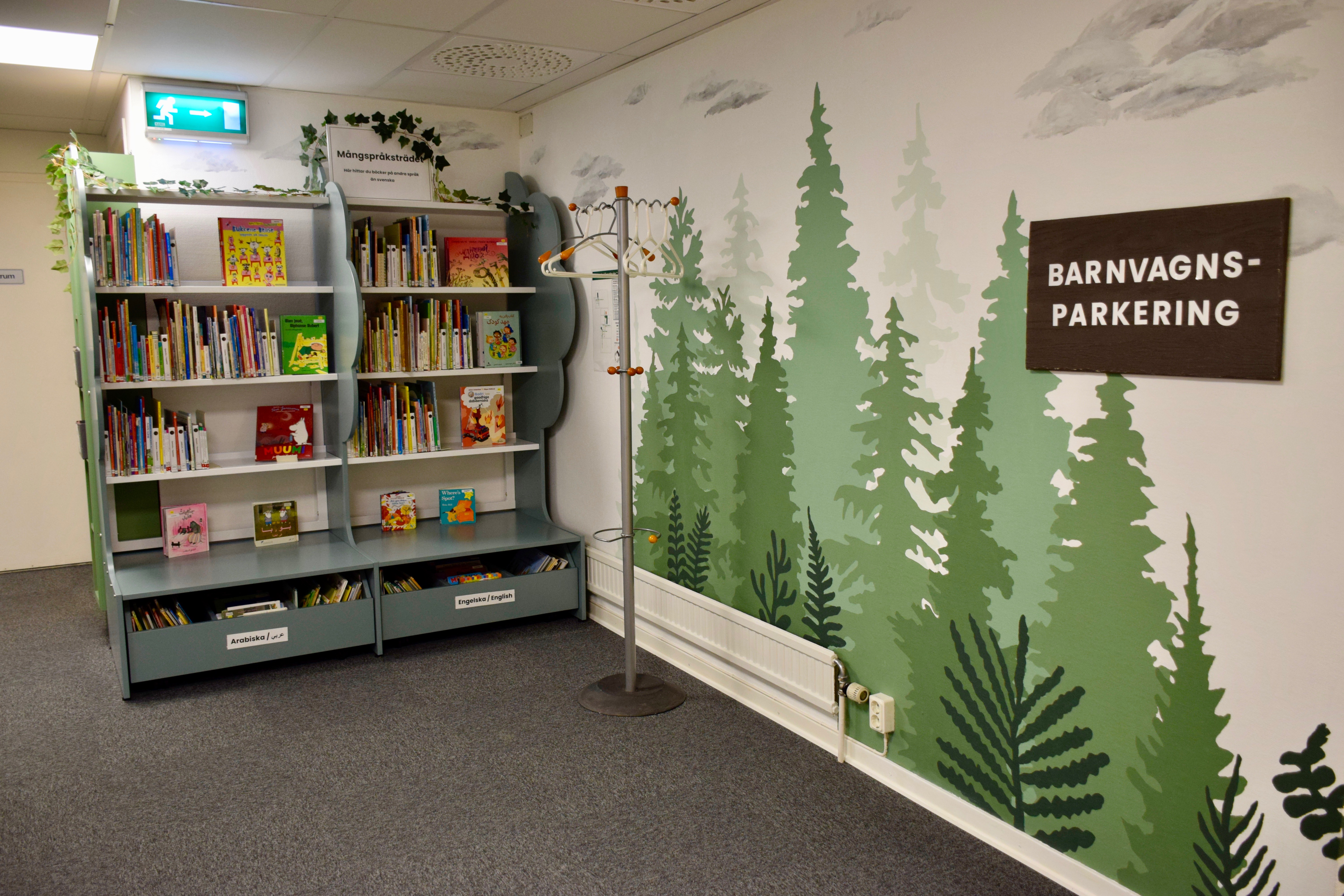 Två gröna bokhyllor fyllda med böcker. På väggen bredvid är granar målade i olika gröna kulörer och på en skylt står det BARNVAGNSPARKERING.