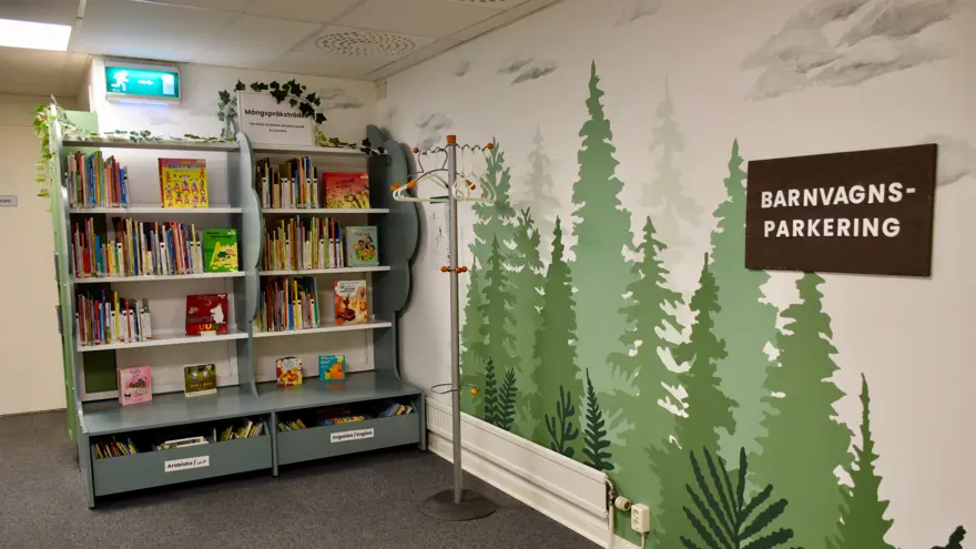 Två gröna bokhyllor fyllda med böcker. På väggen bredvid är granar målade i olika gröna kulörer och på en skylt står det BARNVAGNSPARKERING.