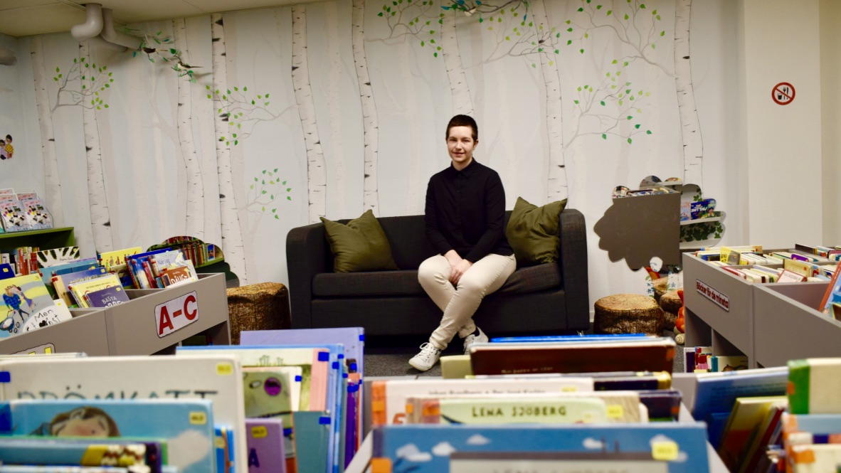 Madeleine Eriksson sitter i en soffa. På väggen bakom är skira björkar målade, i förgrunden finns boklådor. 