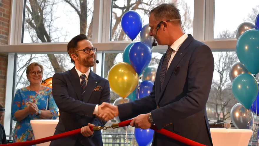 Två män i kostym står på en scen, de skakar hand, tittar på varandra och ler. De håller även i ett tjockt band med guldlänkar som de krokat ihop.