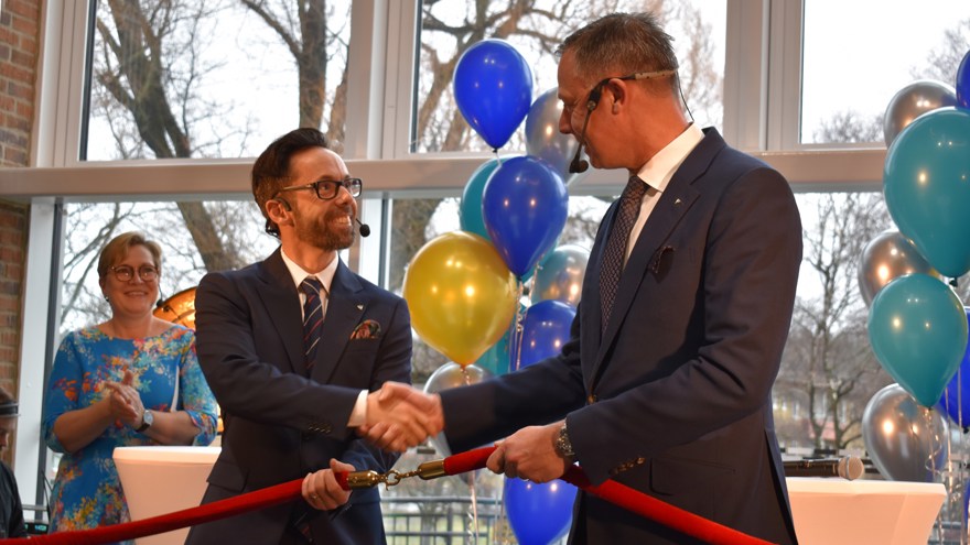 Två män i kostym står på en scen, de skakar hand, tittar på varandra och ler. De håller även i ett tjockt band med guldlänkar som de krokat ihop.