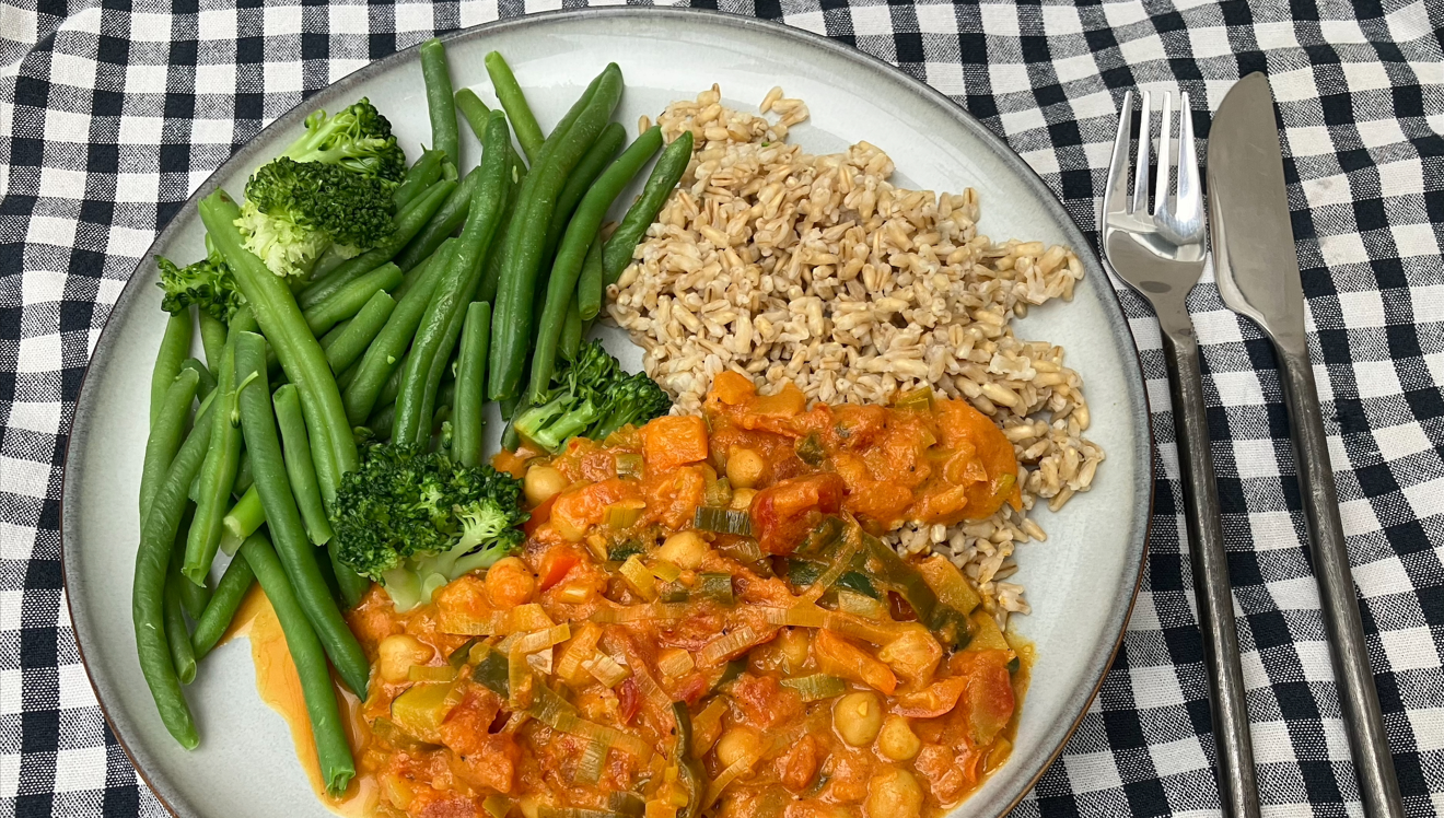 En portion Kikärtsgryta med broccoli och bönor samt havreris på en grå tallrik med bestick och en svrtvit duk