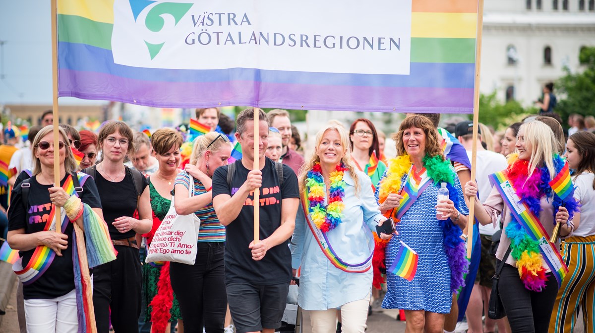 Fotografi taget framifrån. En grupp personer som bär en prideflagga med Västra Götalandsregionens logotyp i samband med West pride