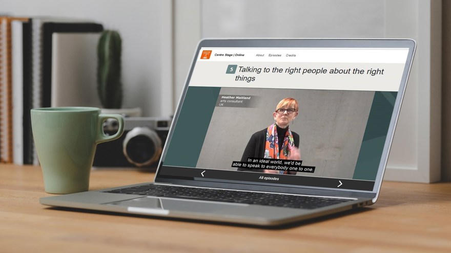 En laptop på ett skrivbord av trä, intill en ljusgrön kaffekopp. På skärmen syns en kvinna i en filmruta, med undertexter och inramat av en turkosfärgad hemsida.