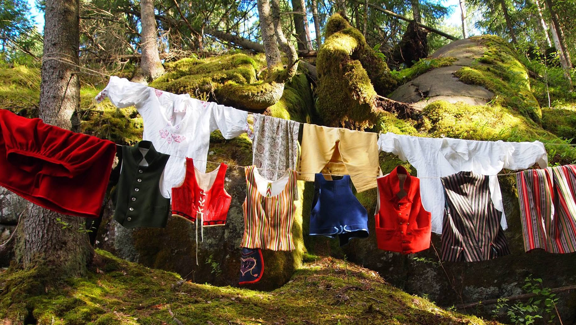 Ett klädstreck framför en träd och mossiga stenar. På sträcket hänger olika däktplagg, vita överdel med broderi, förkläde med tryckt blommönster, gula byxor, vit överdel, röd yllekjol, görn mansväst, kvinnolivstycken i rött, ett randigt och ett blått. Mansvästar en röd och en randig och en randig kjol.