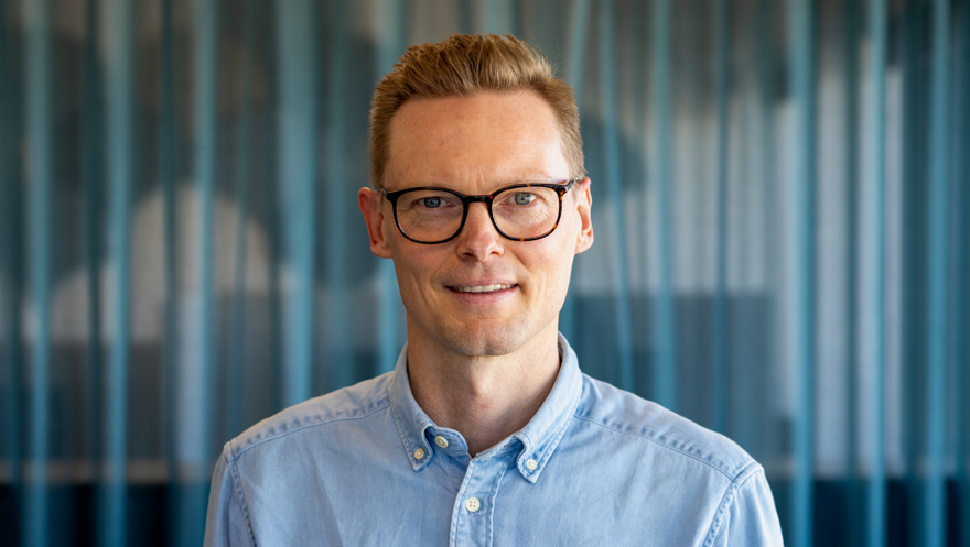 Man i ljusblå skjorta, blont kort hår och glasögon. I bakgrunden ett blågrönt draperi.