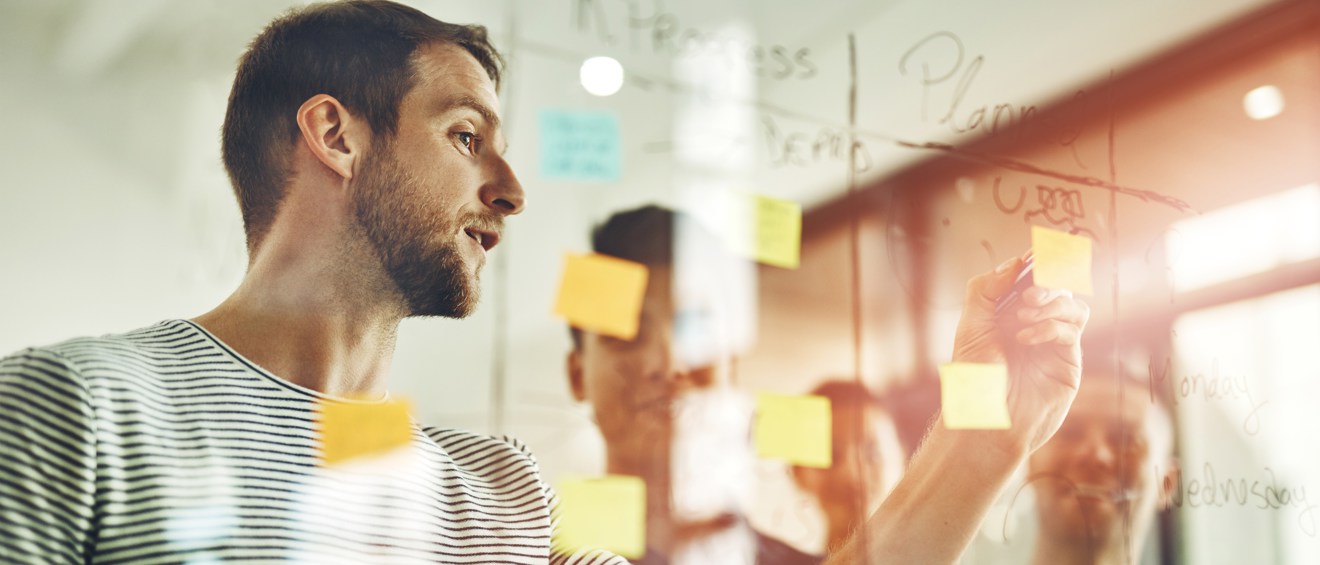 Halvbild som visar en person bakom en glasruta. Glasrutan har flera lappar och anteckningar uppsatta och personen skriver på en av lapparna. I bakgrunden syns tre suddiga personer.