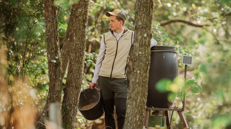 Elev med jaktkläder står bland träd i skogen och håller en hink
