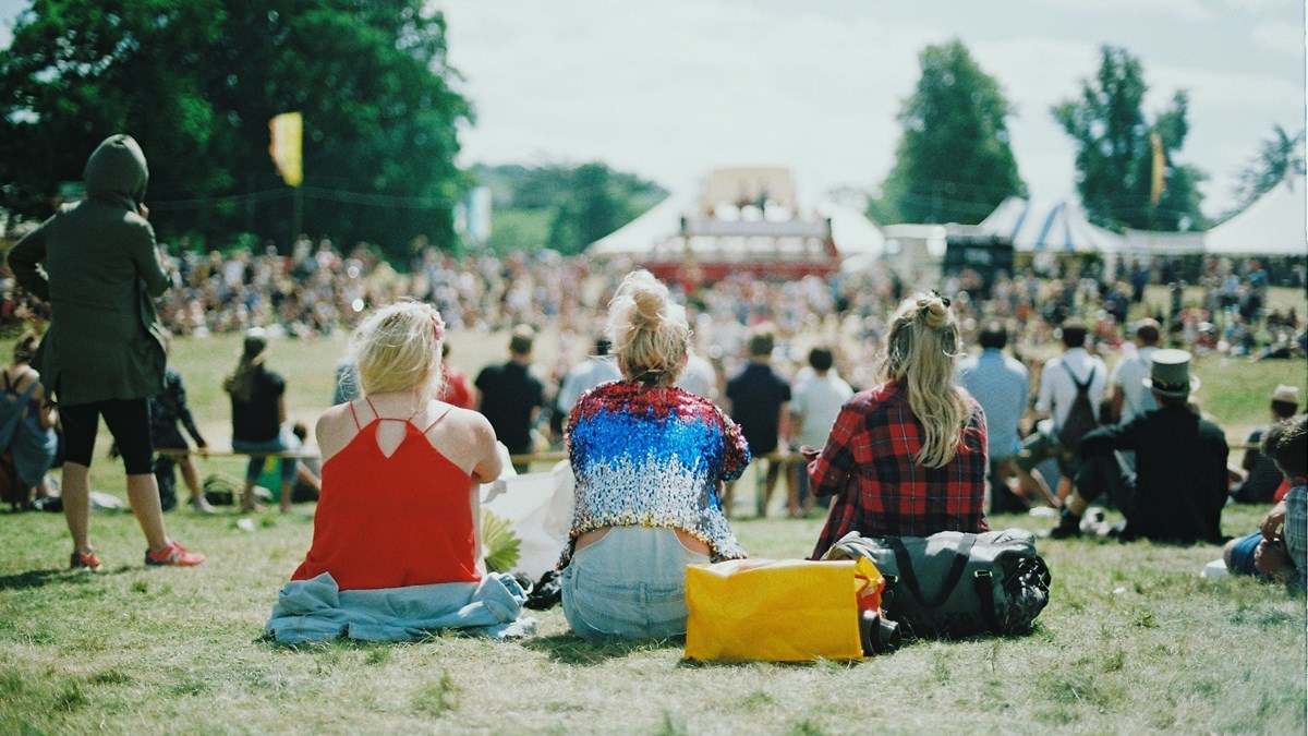 Stor folksamling utomhus på gräsmatta, i bakgrunden evenemangstält dit de riktar uppmärksamheten. I förgrunden tre kvinnor bakifrån, sittandes i gräset med färgglada tröjor och rufsiga ljusa hår. 