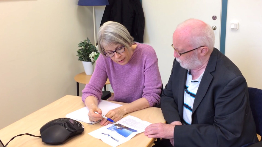 Taltjänsttolken och användaren sitter vid bordet och läser ett reklamblad.