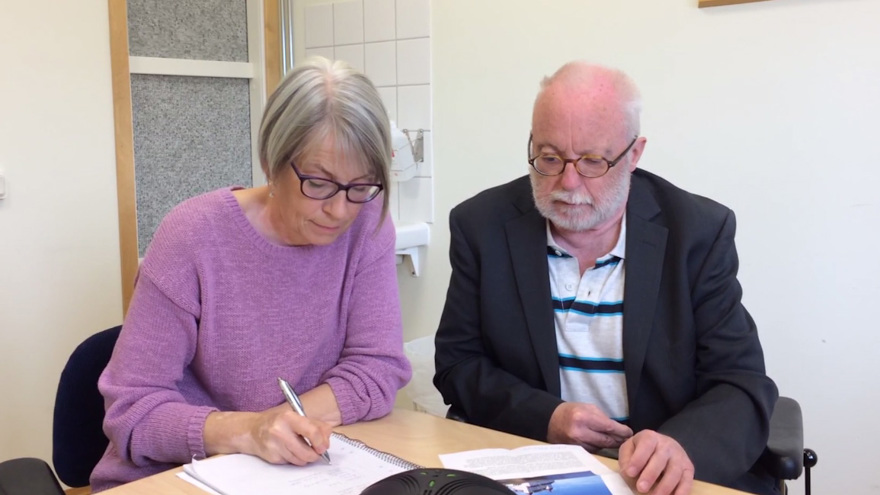 Taltjänsttolken skriver i ett anteckningsblock, användaren sitter bredvid.