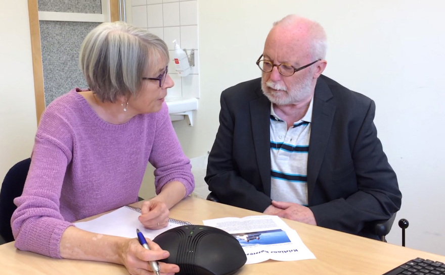 En taltjänsttolk med en penna i handen sitter vid ett bord tillsammans med en taltjänstanvändare.