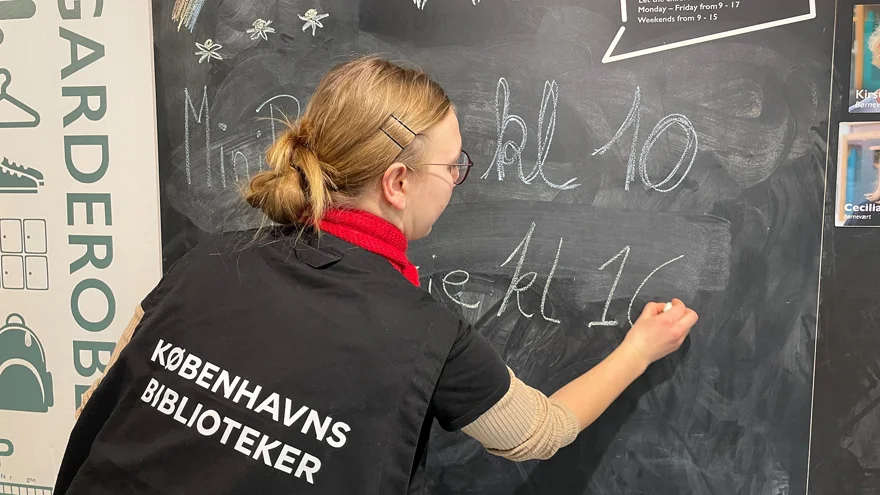 En bibliotekarie skriver dagens program på en svart tavla. Bibliotekarien har en svart väst med texten Köpenhamns bibliotek på ryggen.