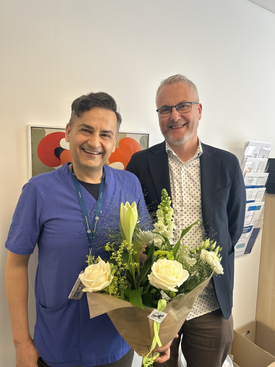 Två leende personer, den ena har blå vårdkläder och håller en stor blombukett.